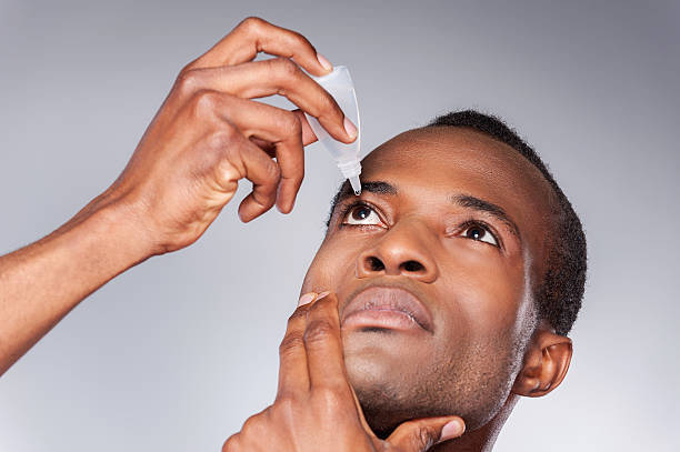 Young African man applying eye drops while standing against grey background